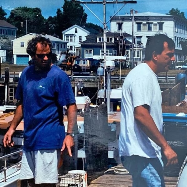 Two men on a dock with houses and a fishing boat in the background.