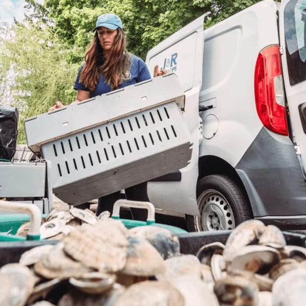 A woman in a J&R Seafood hat and shirt unloads a crate of fresh oysters from a white van.