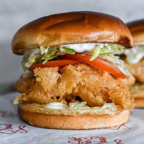 Close-up of a crispy fried fish sandwich with lettuce, tomato, and tartar sauce on a bun.