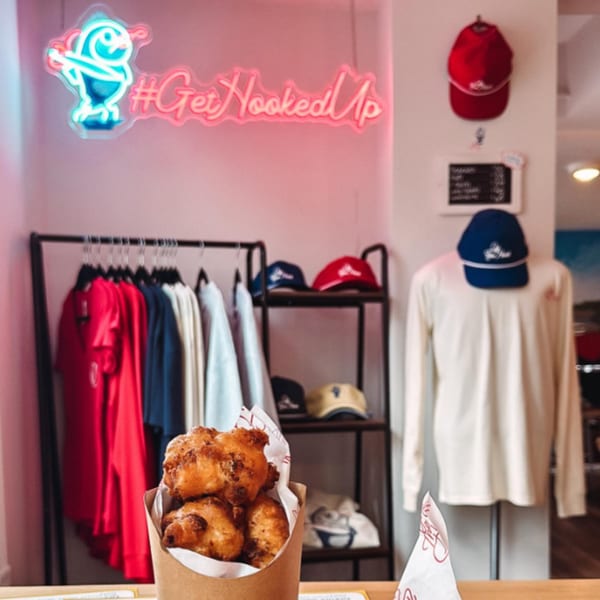 Hushpuppies in a paper cone, with Off The Hook merchandise and a neon sign in the background.