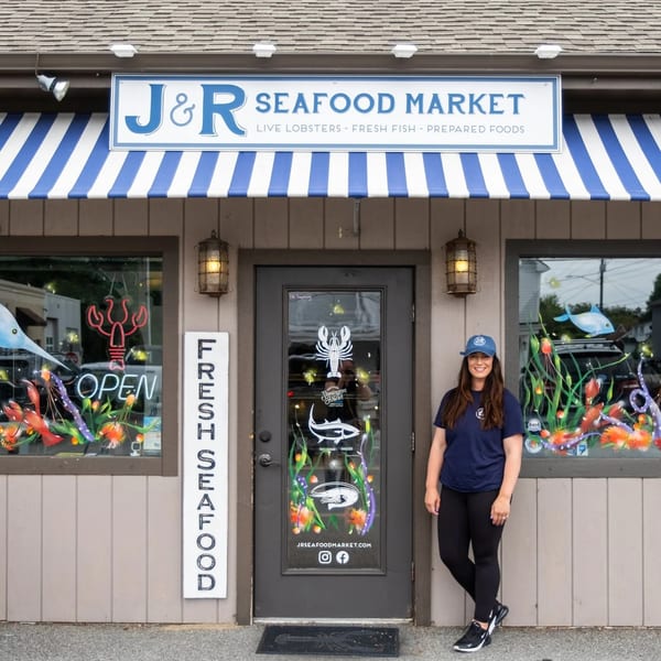 A woman in a blue cap and shirt stands outside J&R Seafood Market with colorful window art.