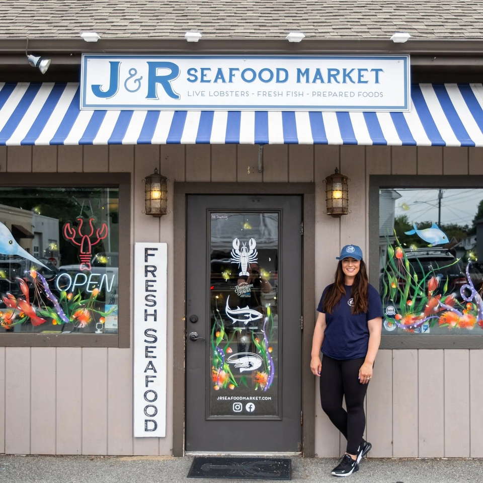 A woman in a blue cap and shirt stands outside J&R Seafood Market with colorful window art.
