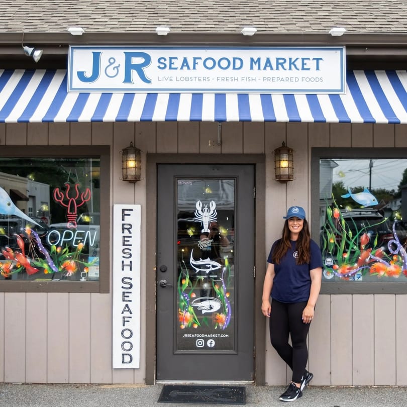 A woman in a blue cap and shirt stands outside J&R Seafood Market with colorful window art.