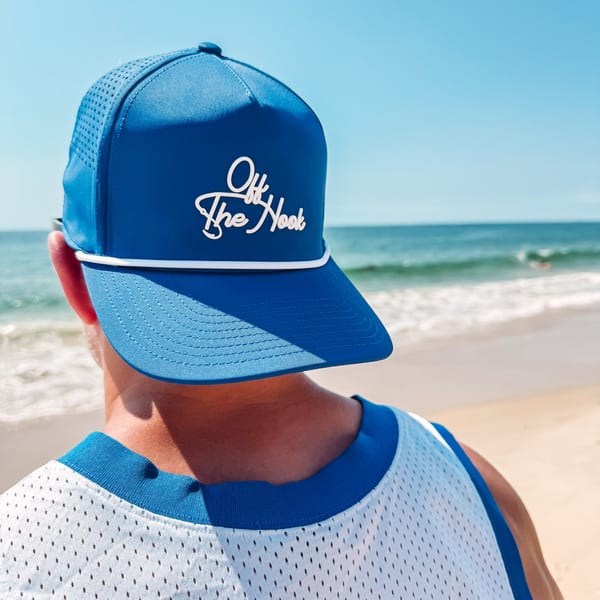 Person wearing a blue "Off The Hook" hat and white jersey, looking at the ocean.