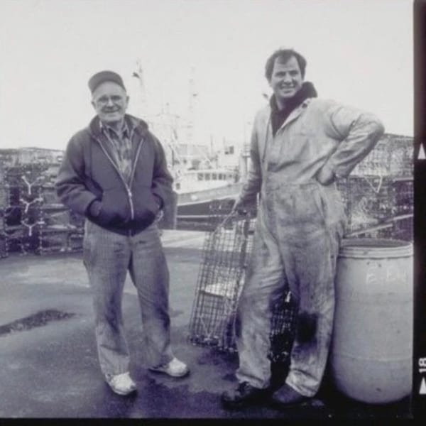 Two men, one holding a lobster trap, stand on a dock with a fishing boat and lobster pots.